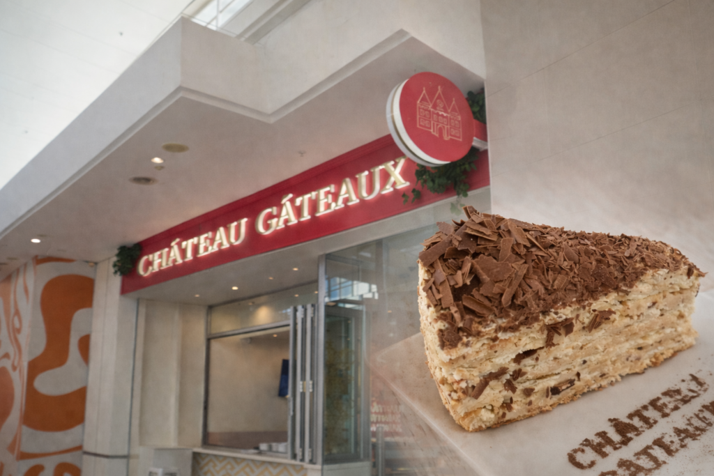 Slice of Mozart cake with layered cream and chocolate shavings in the foreground, with the Château Gâteaux bakery entrance and red signage in the background