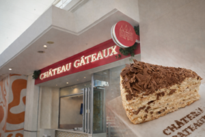 Slice of Mozart cake with layered cream and chocolate shavings in the foreground, with the Château Gâteaux bakery entrance and red signage in the background